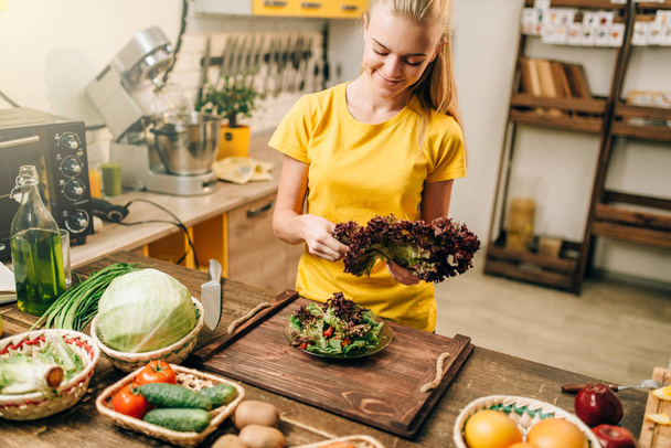 Szczęśliwa młoda kobieta gospodarstwa sałata w kuchni, gotowanie zdrowe jedzenie. Dieta wegetariańska, świeże warzywa i owoce na drewnianym stole - Zdjęcie, obraz