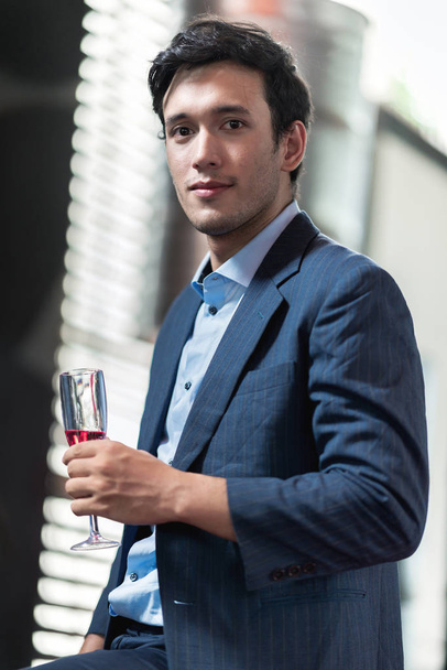 Portrait of young man holding a glass of sparkling rose wine. Handsome chinese single man holding alcoholic beverages with real open air dance club in background. Party concept. - Foto, Imagem