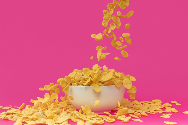 крупным планом вид сладких вкусных кукурузных хлопьев, падающих в белую миску на розовом
  - Фото, изображение