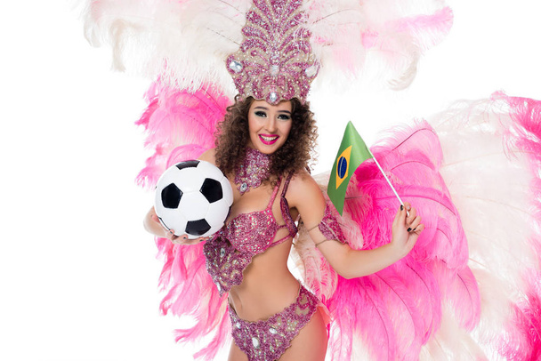 χαμογελαστή γυναίκα στην Αποκριάτικη στολή κρατώντας την μπάλα ποδοσφαίρου και τη βραζιλιάνικη σημαία ενώ βλέπουν τα φωτογραφικών μηχανών, απομονώνονται σε λευκό     - Φωτογραφία, εικόνα