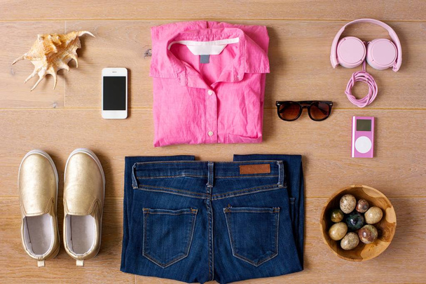 Casual Outfit auf einem hölzernen Hintergrund ausgelegt. Schuhe, Jeans, rosa Hemd, Sonnenbrille, Telefon, pinkfarbener Musikplayer und Kopfhörer. Muschel und eine Schale mit farbigen Steinen als Sidepieces.  - Foto, Bild