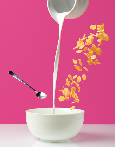 gieten van verse gezonde melk van lokale KRUIK aan kom, lepel en cornflakes op roze     - Foto, afbeelding