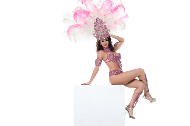 Nuori onnellinen nainen karnevaaliasussa vaaleanpunaisine höyhenineen istuu käsivarsi pään takana eristettynä valkoiseen.
 - Valokuva, kuva