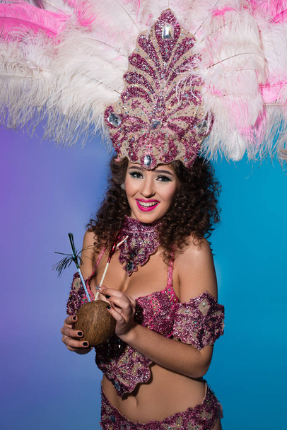 Mulher brilhante em traje de carnaval com penas rosa bebendo de coco inteiro isolado no fundo azul
 - Foto, Imagem