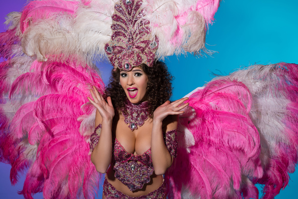 Mujer joven sorprendida en traje de carnaval con plumas rosadas aisladas sobre fondo azul
 - Foto, Imagen