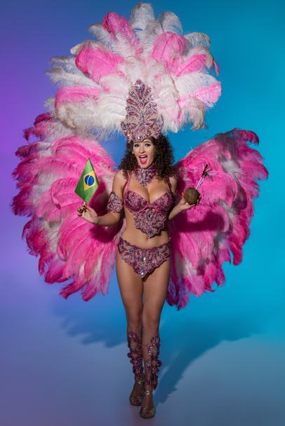 Cheerful woman in carnival costume with pink feathers holding coconut and Brasil flag on blue background - Fotografie, Obrázek