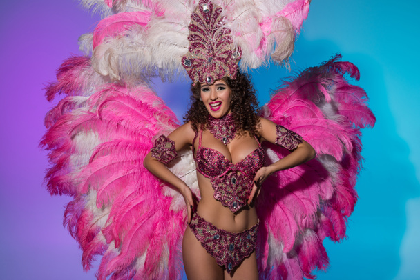 Щаслива молода жінка в карнавальному костюмі з рожевим пір'ям емоційно танцює на синьому фоні
 - Фото, зображення