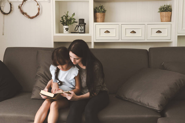 Mother reading a book to daughter in living room together. Family fun time concept. - Foto, imagen