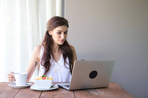Woman having coffee and working outside. Working on laptop during her vacation in a tropical hotel. White female with white dress. Agile. modile office concept. - Фото, изображение