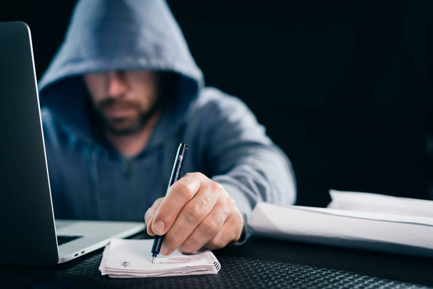 a young guy hacker in a sweatshirt with a hood sits behind a laptop and holds in his hand a pen with a notebook - Photo, image