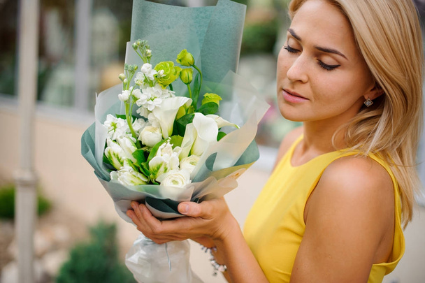 Blonde woman dressed in a yellow dress holding a white and green bouquet of flowers - Fotó, kép