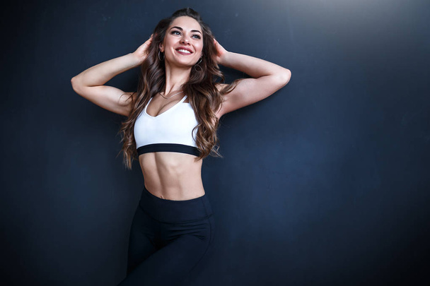 Female fitness model in white top and black leggings is posing in front of a dark blue wall - Fotoğraf, Görsel