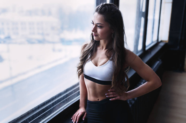 Female fitness model in white top and black leggings is posing near the big windows in studio in morning time - Photo, Image