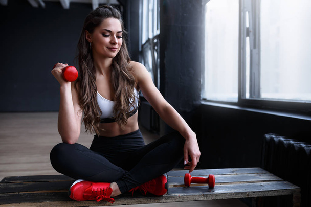 Female fitness model is sitting on bench doing exercises with red dumbbells or relaxing before next set - Photo, Image