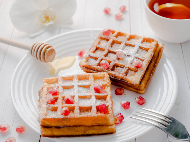 Delicious mouth-watering Viennese waffles with honey and pomegranate seeds on a white plate, light wooden background - Фото, зображення