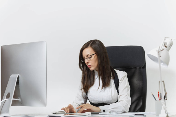 Belle femme d'affaires brune sérieuse et absorbée en costume et lunettes assis au bureau, travaillant à l'ordinateur avec un moniteur moderne avec des documents dans le bureau léger, sur fond blanc
 - Photo, image