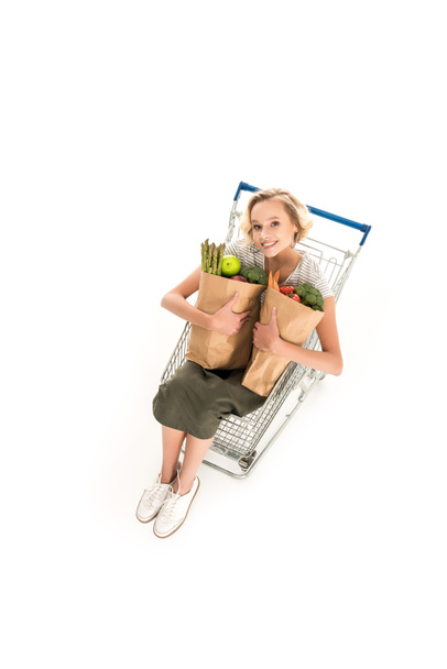 visão de alto ângulo da menina sorrindo para a câmera enquanto segurando sacos de supermercado e sentado no carrinho de compras isolado no branco
 - Foto, Imagem