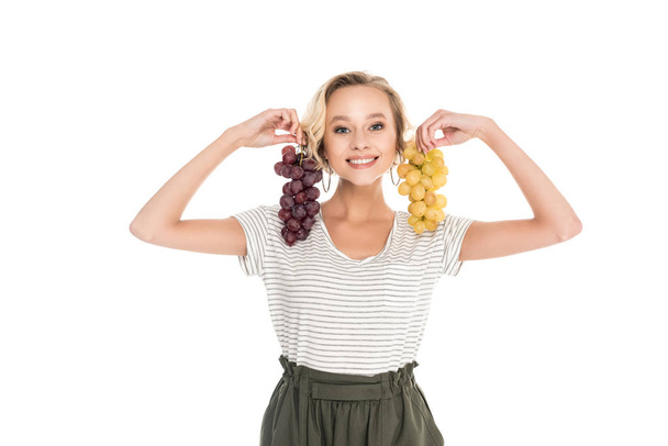 hermosa mujer joven sosteniendo racimos de uvas y sonriendo a la cámara aislada en blanco
 - Foto, Imagen