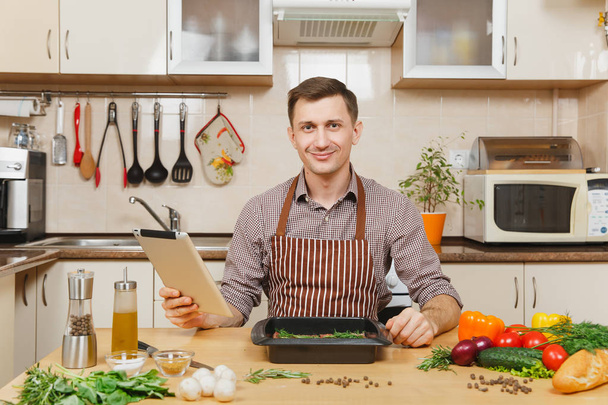 Young European man in apron sitting at table with vegetables, looking for recipe in tablet, cooking at home preparing meat stake from pork, beef or lamb, in light kitchen full of fancy kitchenware. - Photo, Image