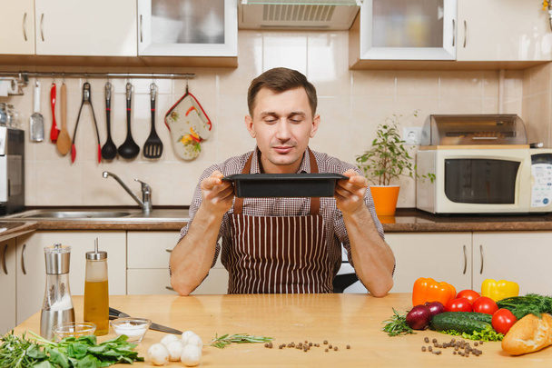 Young man in apron sitting at table with vegetables, inhaling flavor of seasonings, cooking at home, preparing meat stake from beef or lamb, in light kitchen with wooden surface, full of kitchenware. - Photo, Image