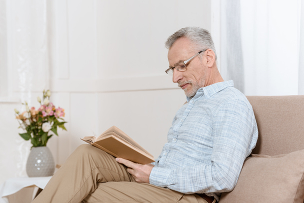 Senior man reading book on sofa in cozy room - Valokuva, kuva