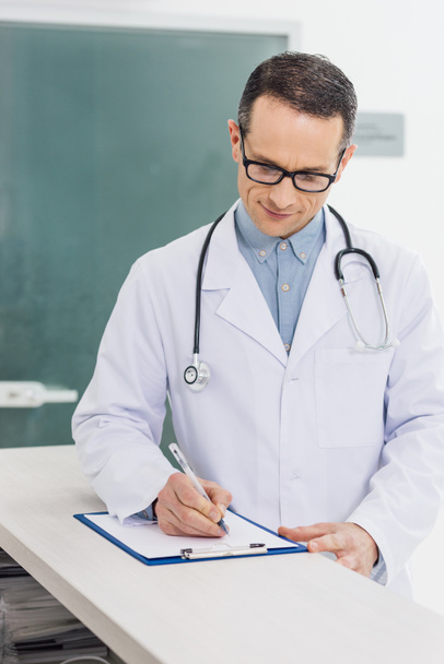lachende arts in witte jas met stethoscoop schrijven diagnose in het ziekenhuis - Foto, afbeelding