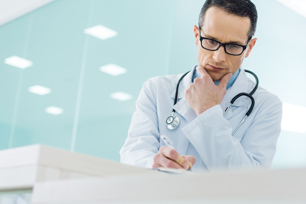 przemyślane lekarz w białym płaszczu z stetoskop pisania diagnozy w szpitalu - Zdjęcie, obraz