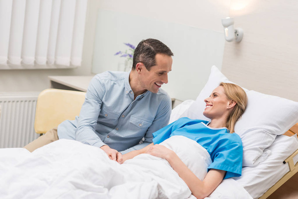 smiling husband sitting at wife bed in hospital - Foto, afbeelding