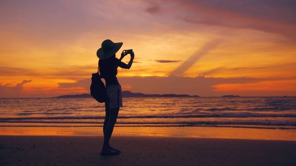 Siluetti nuori turisti nainen hattu ottaa valokuvan matkapuhelin auringonlaskun aikana valtameren rannalla
 - Valokuva, kuva