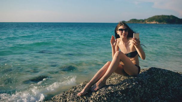 Молодая счастливая женщина чатится с друзьями по интернету, используя смартфон на пляже океана
 - Фото, изображение