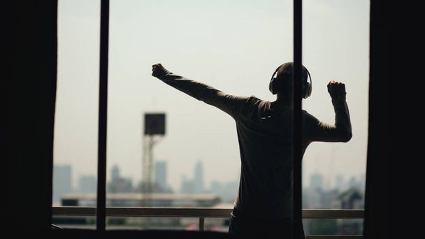 Silhouette of young man dancing ad listening music in wireles headphones stand on hotel room balcony - Photo, image