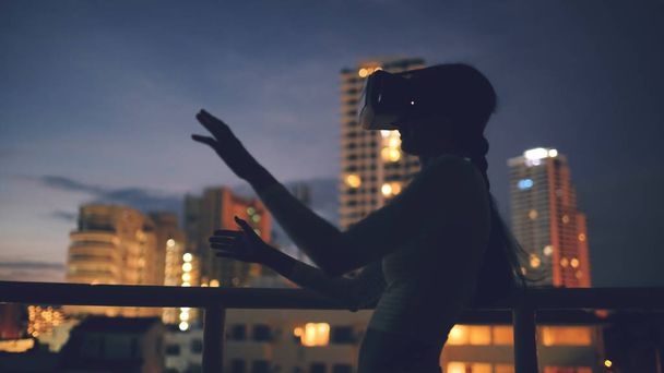 Νεαρή γυναίκα στη βεράντα τελευταίου ορόφου χρησιμοποιώντας σετ κεφαλής εικονικής πραγματικότητας και έχοντας εμπειρία εικονικής πραγματικότητας, το βράδυ - Φωτογραφία, εικόνα