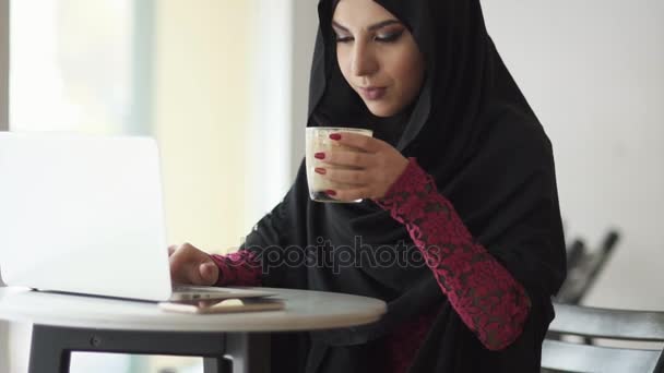 Young muslim woman in hijab sitting in cafe drinking a cup of coffee and using her laptop. She is searching for something in internet. Studying or working. Slowmotion shot - Záběry, video