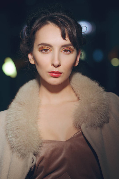 Portrait of beautiful girl in coat with fur collar posing in dark room with lights on background - Fotó, kép
