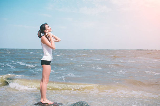 portrait of young sporty woman in headphones relaxing while sitting near ocean in summer , attractive female listening favorite music during recreation time in weekend. - 写真・画像