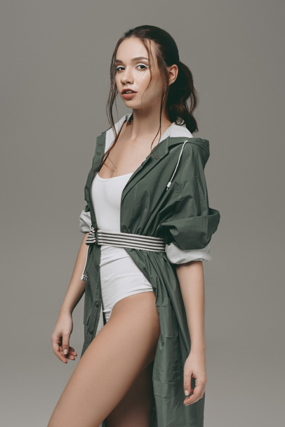 attractive young girl posing in bodysuit and raincoat, isolated on grey - Photo, Image