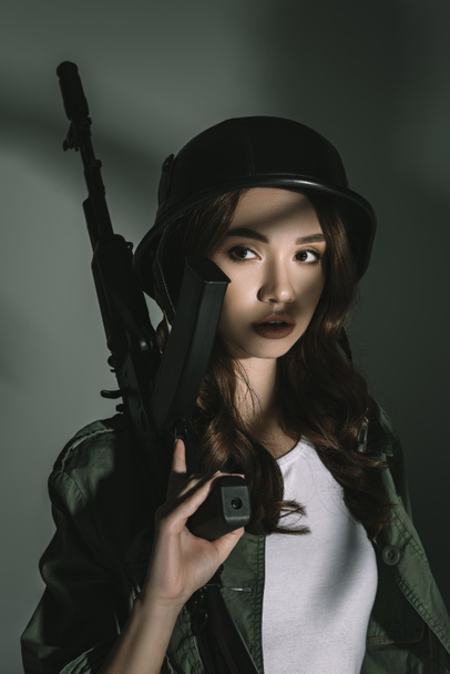 όμορφη κοπέλα ποζάρει στο στρατιωτικό κράνος με το τουφέκι, στο γκρι με σκιές - Φωτογραφία, εικόνα