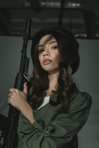 beautiful girl posing in military helmet with rifle, on grey with shadows - Photo, Image