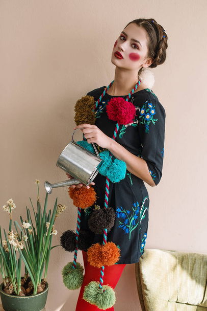 Fashionable beautiful brunette girl in a jumpsuit with floral embroidery with color makeup: red cheeks and lips having fun, watering flowers. Granny chic style. Retouched portrait - 写真・画像
