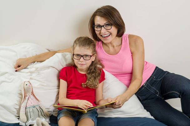 Happy woman with her daughter child, reading together a book at home. Mom and daughter wearing glasses. - Zdjęcie, obraz