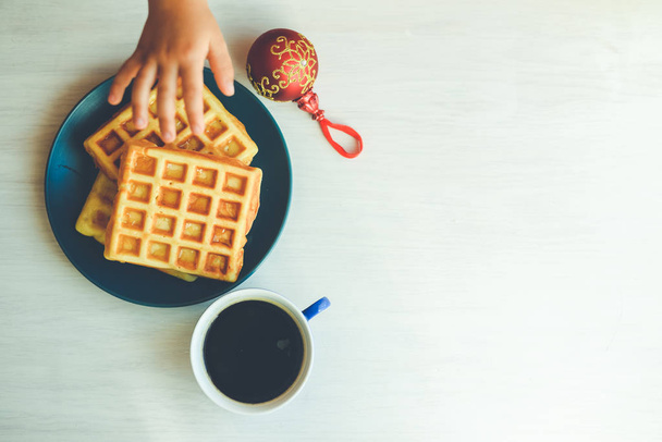 Closeup of child hand and baked waffles on light table background. Top side view on tasty natural decoration. Xmas classic lifestyle. Winter style delicious pastries. - 写真・画像
