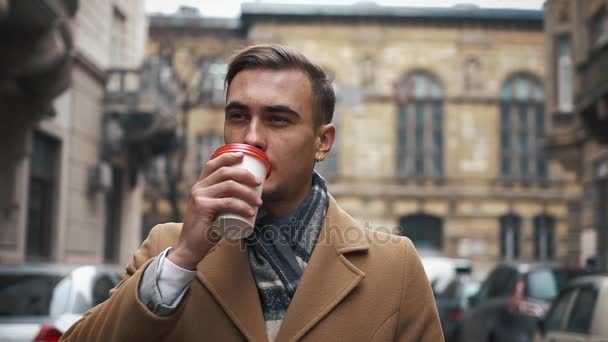 ein junger Geschäftsmann in einem Mantel, der bei bewölktem Wetter die Straße hinuntergeht und Kaffee trinkt. Zeitlupe - Filmmaterial, Video
