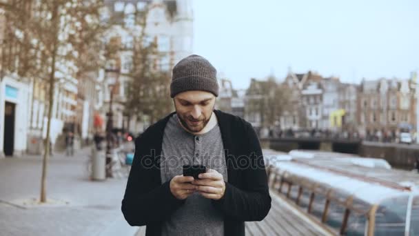 4k kaukasischen Touristen Mann zu Fuß mit Smartphone. lässige gutaussehende bärtige lächelnde erwachsene männliche SMS auf der Straße. - Filmmaterial, Video