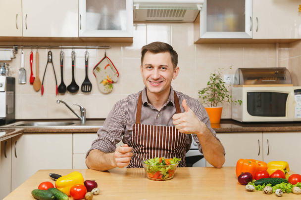 attraktiver kaukasischer junger Mann in Schürze, braunes Hemd am Tisch mit Gemüsesalat in Schüssel, Daumen hoch in heller Küche. Diätkonzept. Gesunder Lebensstil. Kochen zu Hause. Essen zubereiten - Foto, Bild