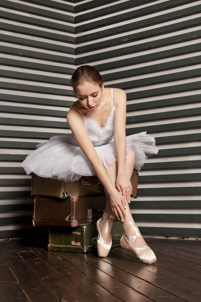 sie ist eine schöne junge Ballerina in einem weißen klassischen Kleid, die auf einem Stapel Retro-Koffer auf einem schwarz-weiß gestreiften Hintergrund sitzt - Foto, Bild