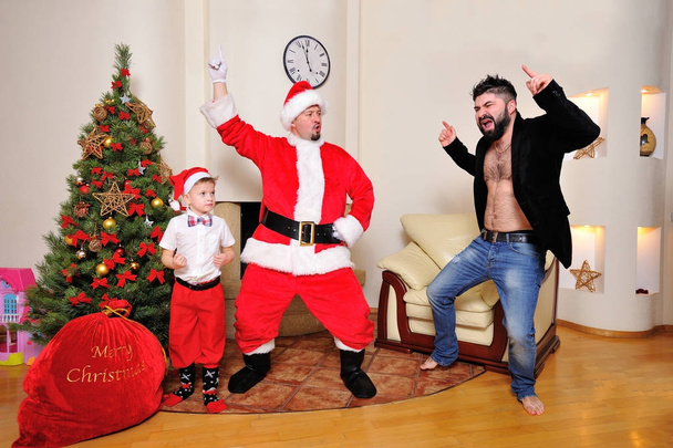 Christmas tree, gift bag, fireplace - men dressed as Santa, boy in red pants and high socks and a hipster in velours jacket on bare torso dancing, play the fool. Looking crazy - Photo, Image