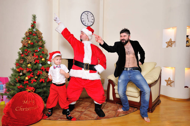 Vánoční strom, taštička, krb - mužů oblečený jako Santa, chlapče a v červených kalhotách a vysoké ponožky a bokové v velur bundu na holé torzo tančit, hrát na blázna. Hledáte blázen - Fotografie, Obrázek
