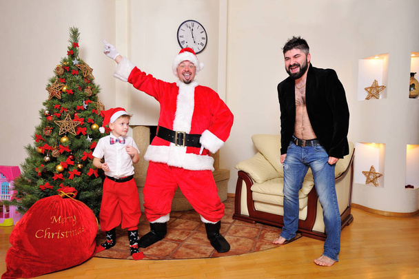 Χριστουγεννιάτικο δέντρο, τσάντα δώρο, τζάκι - άνδρες ντυμένοι ως Σάντα, αγόρι με κόκκινο παντελόνι και κάλτσες και ένα hipster σε βελουτέ σακάκι γυμνού κορμού χορού, παίζουν ο ανόητος. Ψάχνουν τρελό - Φωτογραφία, εικόνα