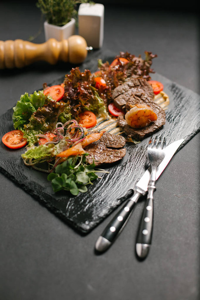 Gegrild rundvlees geserveerd met verse groenten en sla bladeren op een zwart bord - Foto, afbeelding