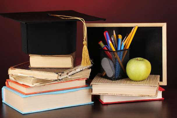 Books and magister cap against school board on wooden table on red background - Foto, afbeelding
